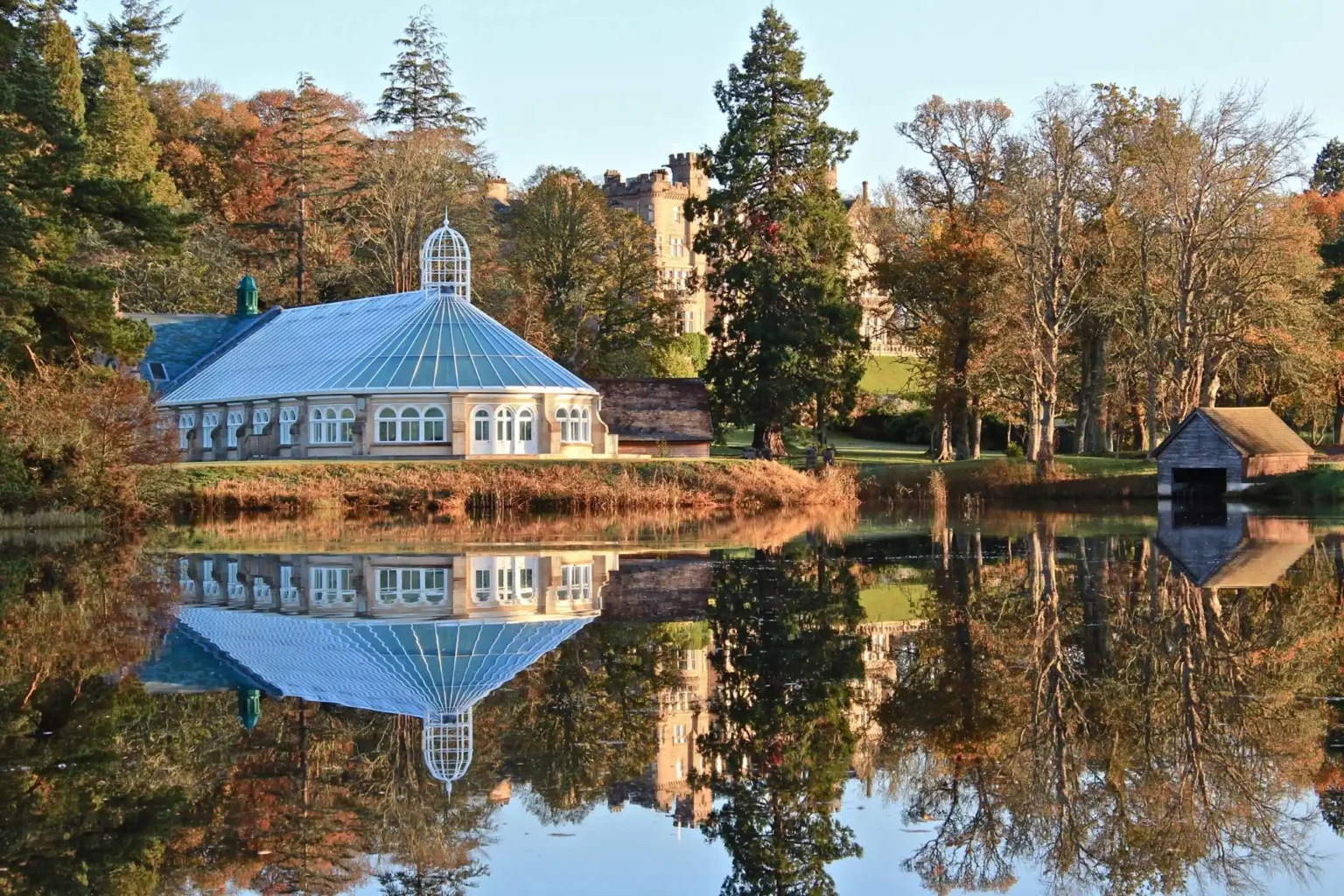 Glasshouse by a lake with autumn trees and reflections in serene water.