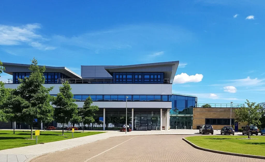 A modern veterinary school building with a striking front elevation, showcasing a combination of reflective glass, clean white panels, and natural stone cladding. The entrance is accented with blue window frames, and the facility is surrounded by lush green landscaping and clear blue skies.