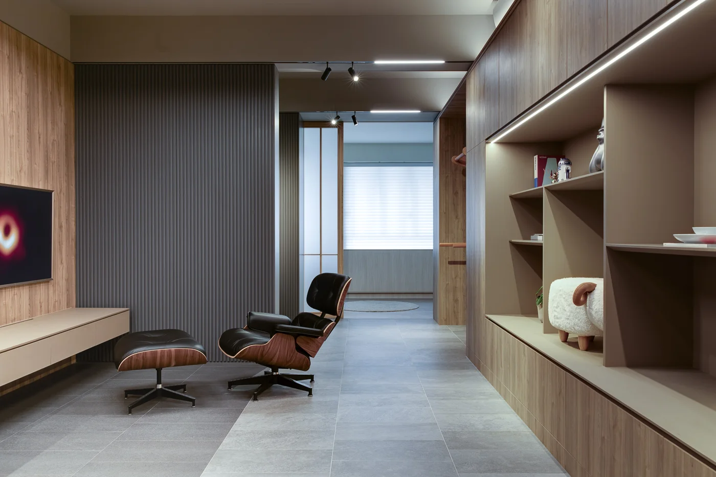 Elegant modern office designed by Rebel Arkitekt, showcasing a luxurious Eames Lounge Chair, sleek built-in shelving with curated decor, and contrasting textured wall panels.