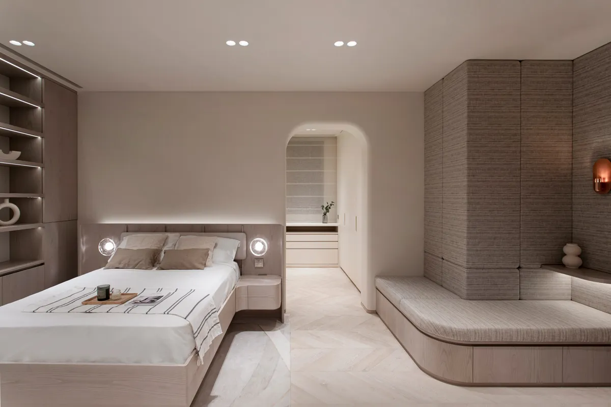 Minimalist bedroom with a queen-sized bed, soft lighting, and an en suite seating area. Modern bookshelves flank the bed, enhancing the clean, serene atmosphere.