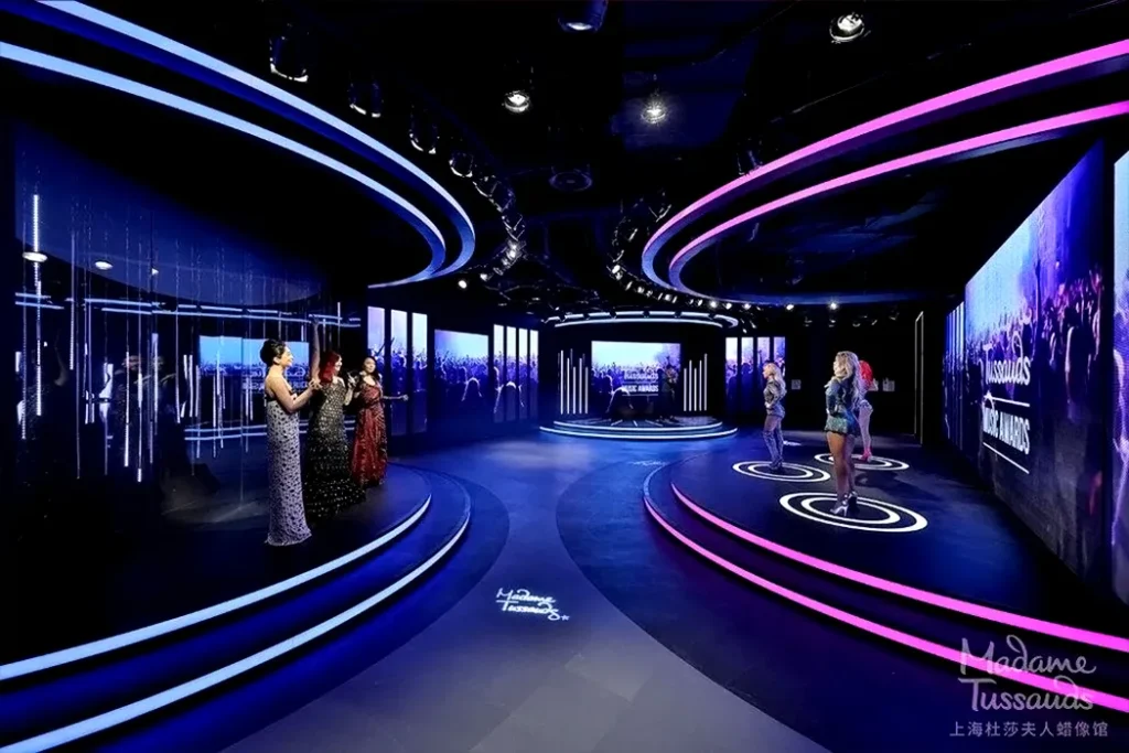 A 360-degree stage with neon blue and pink lights at Madame Tussauds showcases wax figures of iconic musicians in various poses, creating an immersive music-themed environment.