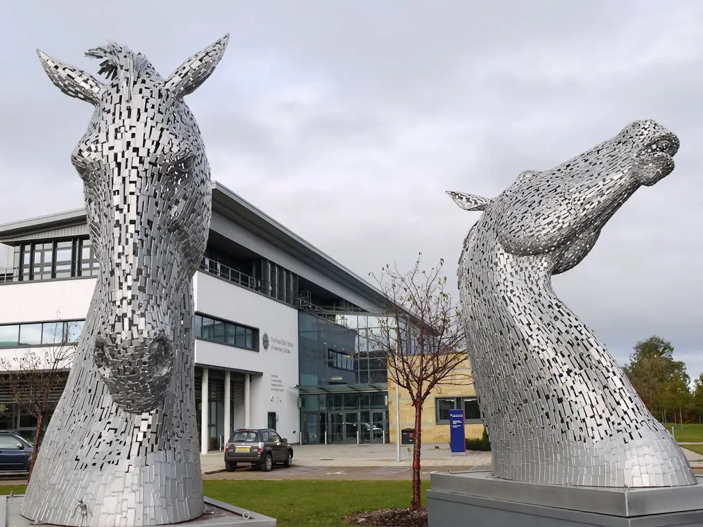 A pair of intricate metallic horse head sculptures in front of the new veterinary school building, with the detailed structure of the building in the background, under a partly cloudy sky