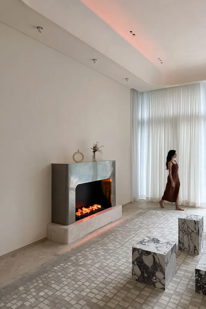 Spacious room featuring a decorative fireplace, sheer draped windows, and marble cube stools. A figure walks along the tiled floor, bathed in natural light.