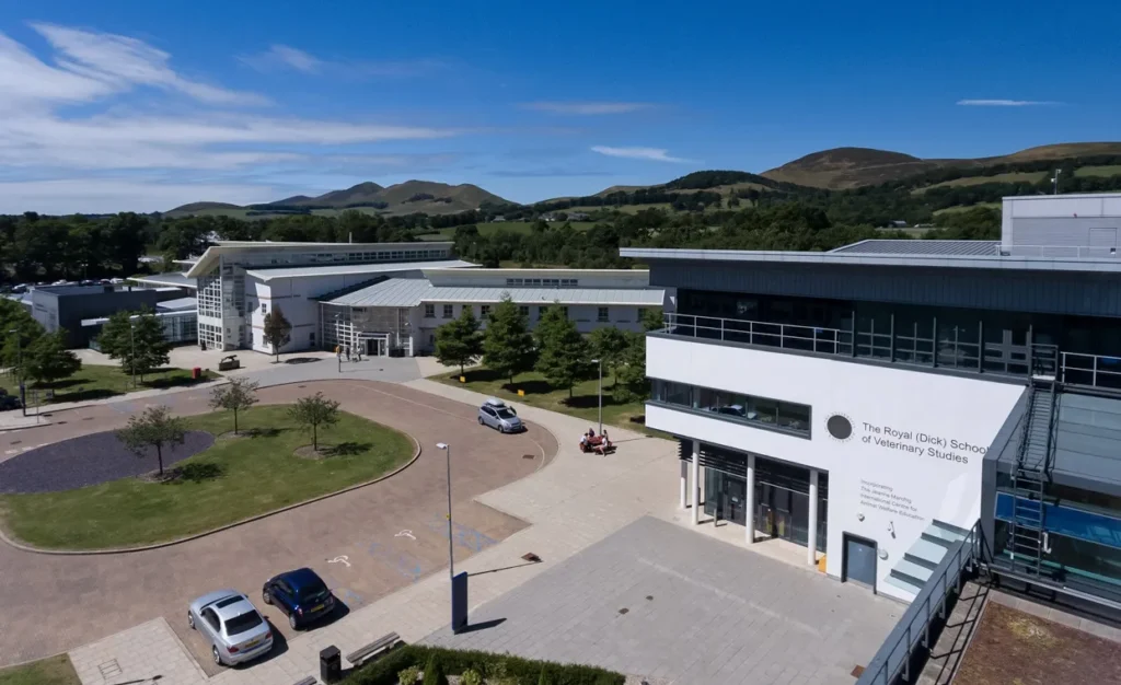 Aerial view of The Royal (Dick) School of Veterinary Studies with a white, modern building in the foreground, featuring large glass windows, a flat roof, and clear signage. The facility is surrounded by well-maintained lawns and parking areas with a few cars and a seating area. In the background, the landscape features rolling green hills under a clear blue sky with a few wispy clouds.