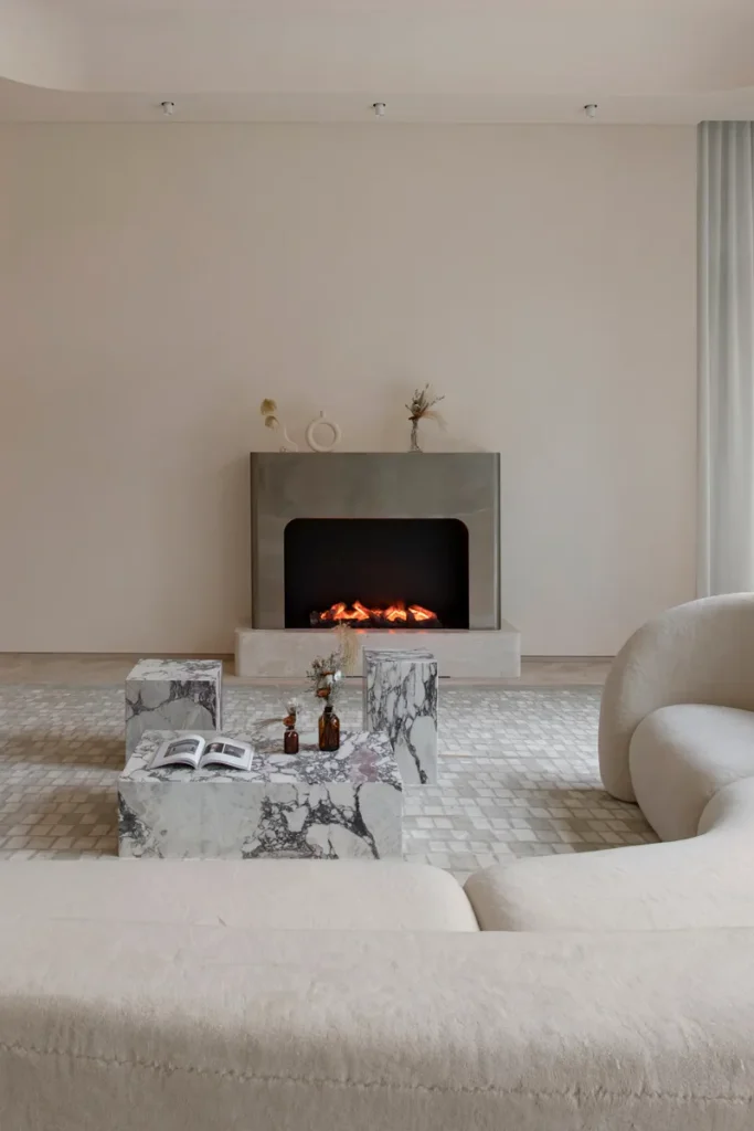 Elegant lounge with a soft-toned curved sofa, a modern fireplace, and accentuated with marble cube side tables against sheer curtains and ambient ceiling lighting.