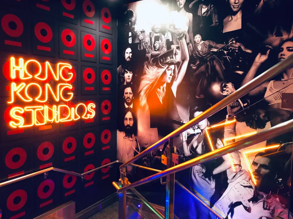 The "Hong Kong Studios" neon sign lights up a wall featuring a collage of iconic music stars at Madame Tussauds, adding a vibrant touch to the themed area.