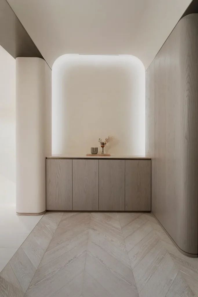 A minimalist hallway with curved walls, herringbone wood flooring, and a built-in light wooden cabinet topped with a simple vase, combining modern design with subtle elegance.