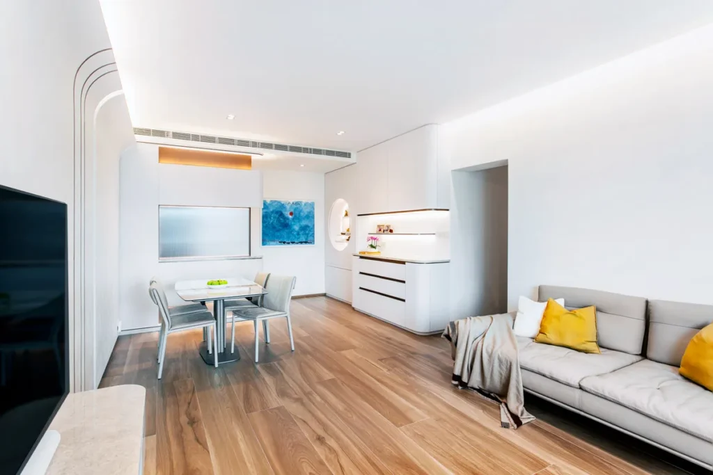 Contemporary living space with a feature curved wall and a glass foyer cabinet with brass accents, designed by Rebel Arkitekt, complementing the wooden flooring and a cozy grey sofa with yellow throw pillows against a backdrop of serene white walls.