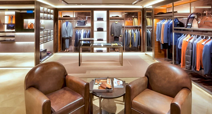 Men's clothing store interior featuring leather chairs, wooden shelving with suits and accessories, and ambient lighting.