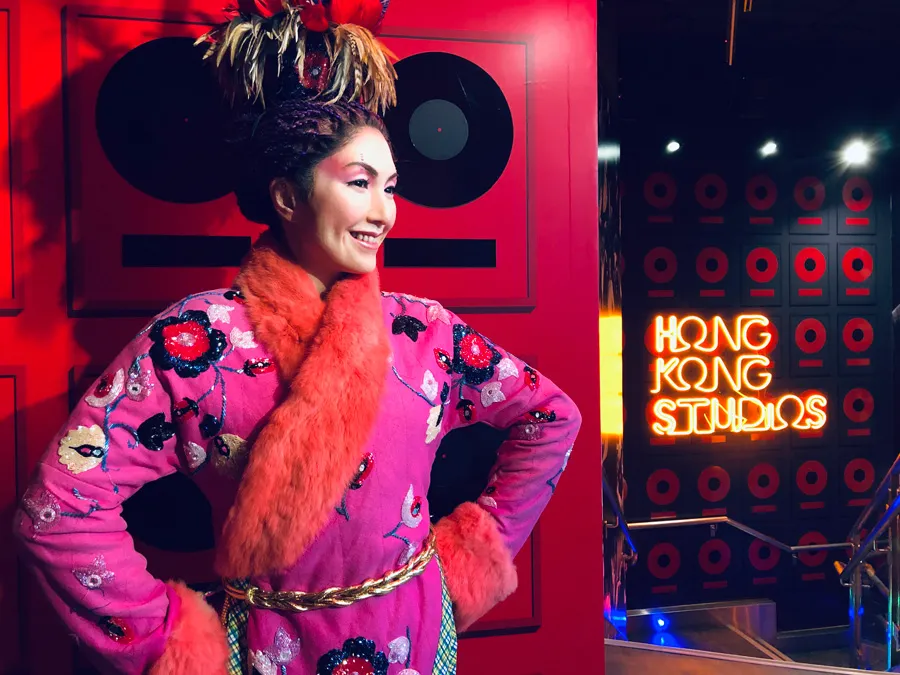 A wax figure clad in a vibrant traditional costume stands before a backdrop of red walls and speakers, with the "Hong Kong Studios" neon sign glowing warmly nearby.