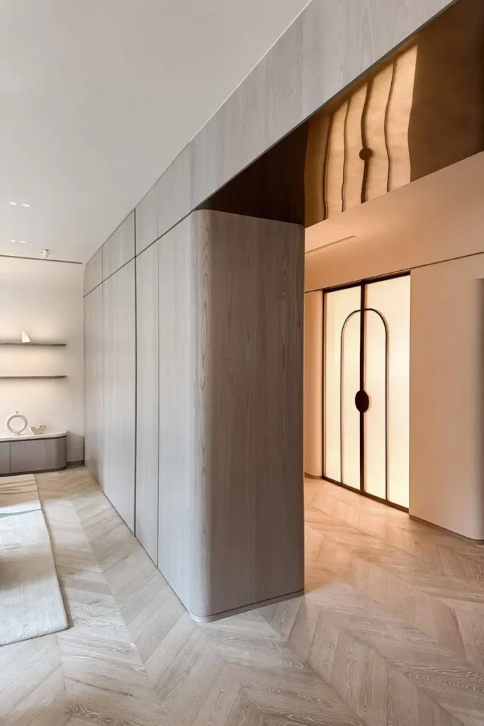 Modern hallway featuring sleek wood paneling and backlit lift doors with a circular design, complemented by herringbone-patterned flooring and warm ambient lighting.