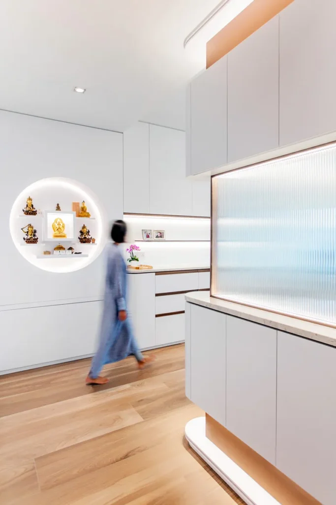 Minimalist entryway crafted by Rebel Arkitekt, featuring a brass-topped glass foyer cabinet, with the founding director in motion, accentuating the flow of the space and the spiritual alcove illuminated in the background.