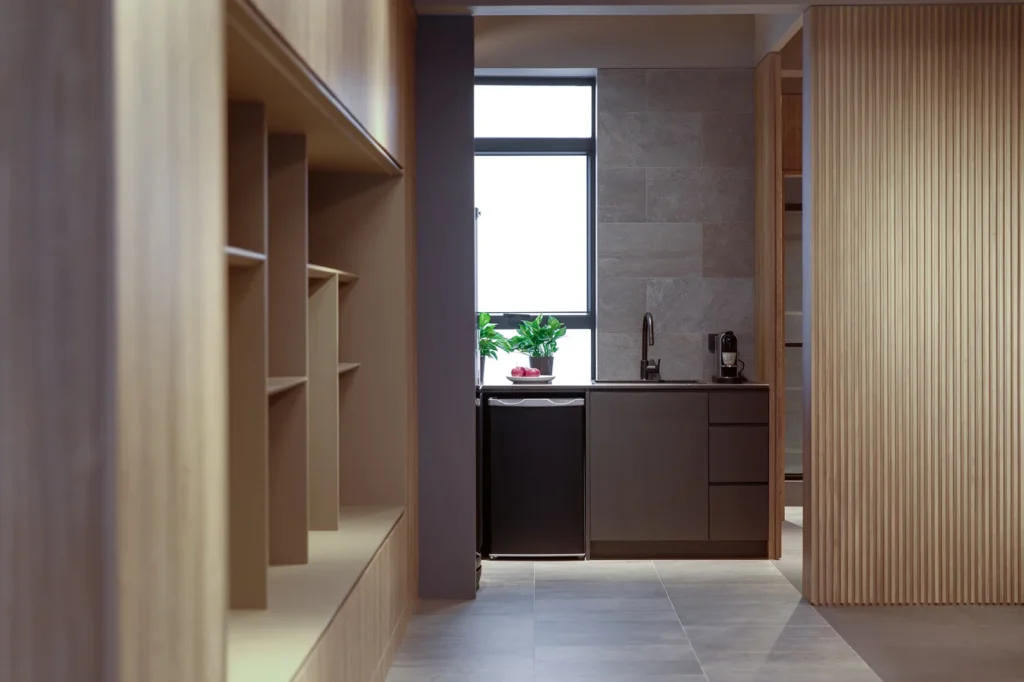 Contemporary office kitchenette and bookshelf design by Rebel Arkitekt featuring sleek cabinetry and strategic lighting for a functional, modern workspace.