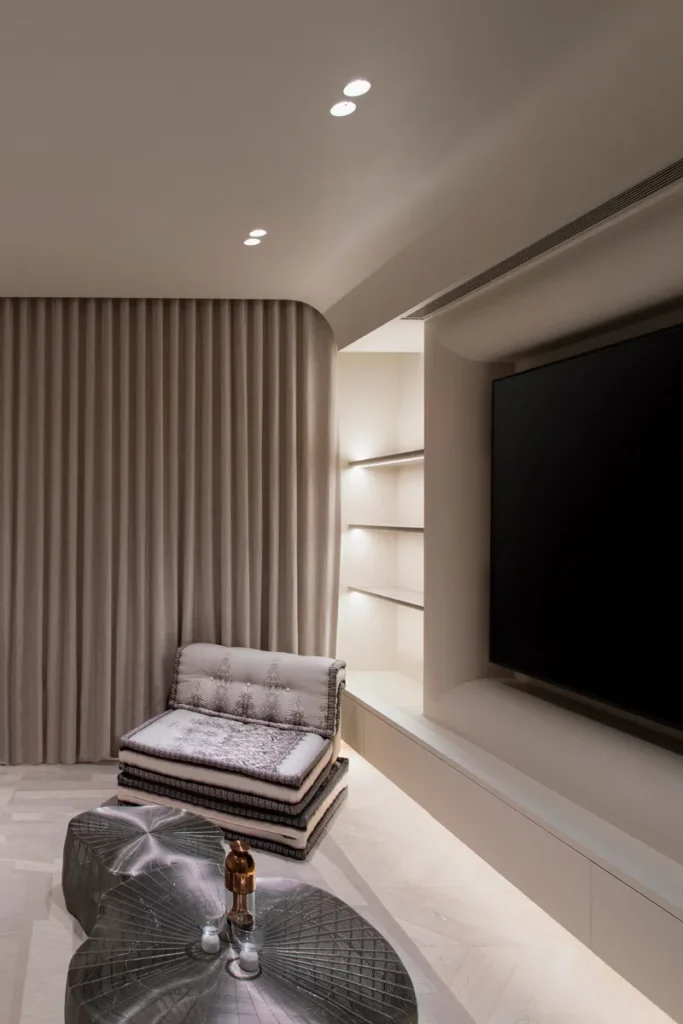 Elegant entertainment space in a luxury suite with soft recessed lighting, featuring a large flat-screen TV, plush drapery, and illuminated shelving, complemented by a sculptural metallic coffee table.