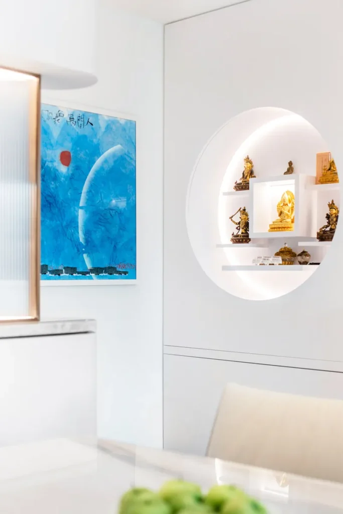 Artfully arranged interior by Rebel Arkitekt, where a blue-toned abstract painting complements the white walls and bespoke round shelving that elegantly displays spiritual figurines, harmonizing modern design with traditional elements.