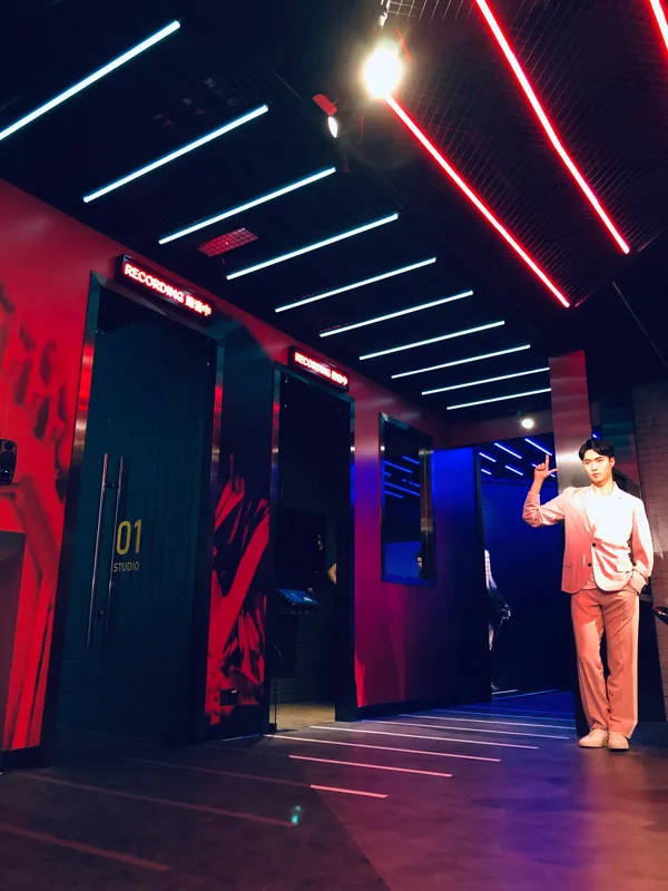 A dimly lit corridor with neon blue and red lighting overhead leads to recording studios, featuring a wax figure in a pink suit gesturing towards one of the doors.