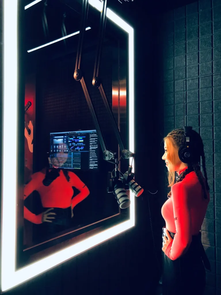 The image displays a recording studio booth illuminated by white neon lights, with a woman wearing a red top and headphones, standing before a microphone, reflected in the glass.