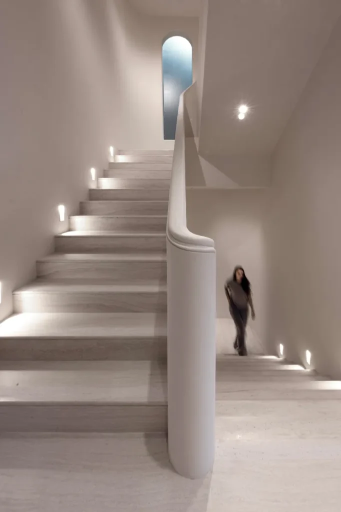 An ethereal staircase in a modern, minimalist interior. The stairs ascend in a soft, sweeping curve, flanked by a slender, sculptural column.