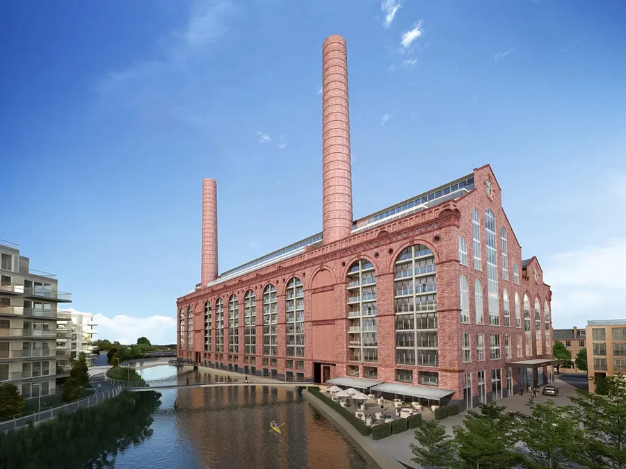 Renovated Lots Road Power Station by the creek, transformed into a residential building with classic red brick architecture and tall chimneys, featuring modern glass windows, adjacent to contemporary buildings, with a serene waterside promenade and outdoor café seating.