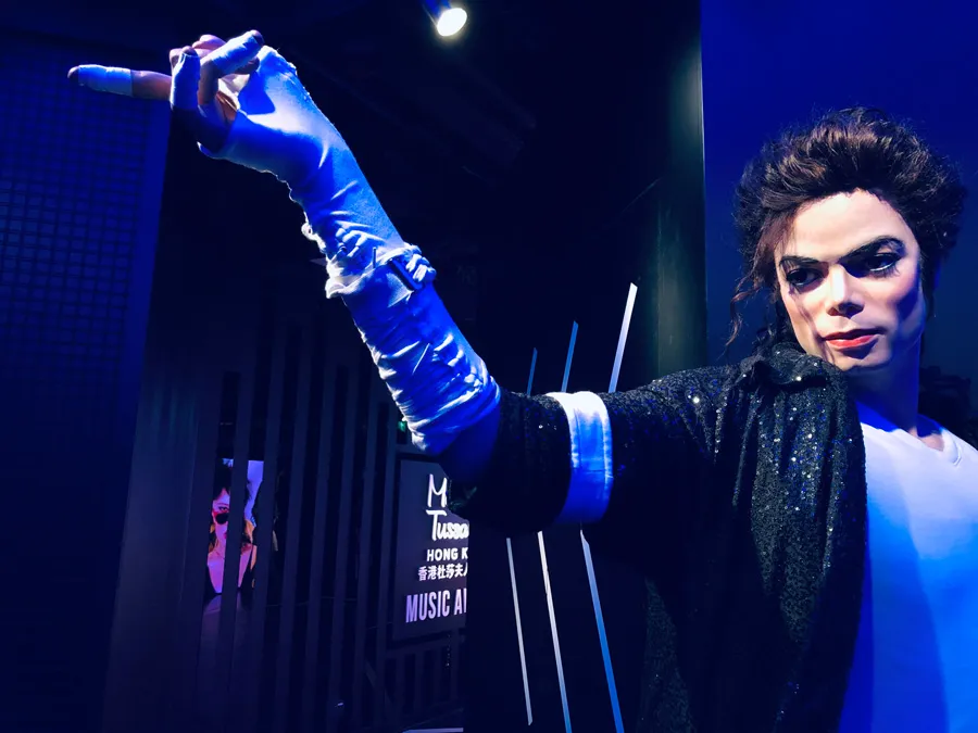 A wax figure of Michael Jackson with dramatic makeup and a sparkling jacket is striking a pose under blue stage lighting.