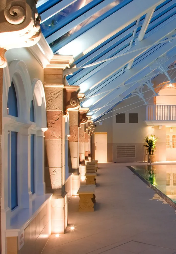 Illuminated poolside corridor with arched windows and ornate pillars under a glass roof.