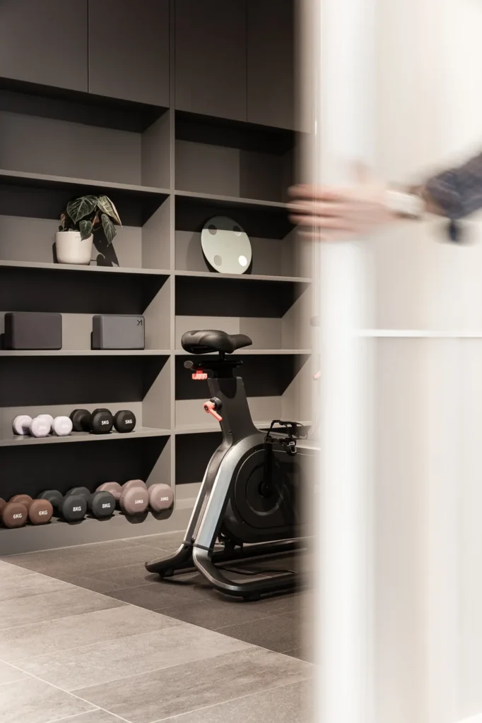 A sleek stationary bike and a set of weights occupy a corner of the Rebel Arkitekt-designed office, promoting health with a sophisticated, built-in black shelving unit that doubles as a stylish, functional display for workout equipment.