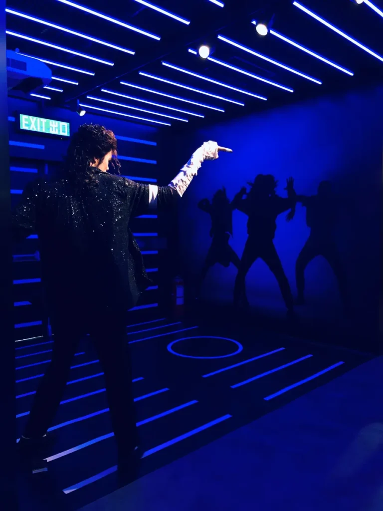 A wax figure of Michael Jackson points forward in a neon-lit room with silhouetted dancers and an 'EXIT' sign above.