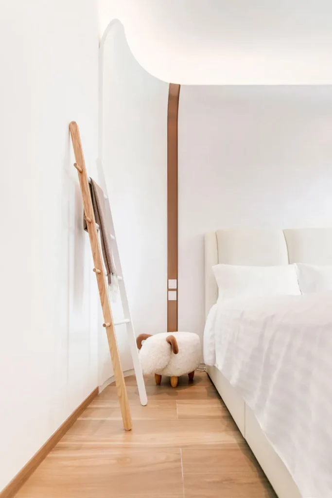 Master bedroom corner with a creative touch by Rebel Arkitekt, featuring a Scandinavian-style clothes rack and whimsical sheep stool, alongside the smooth lines of the bed and an arched bronze detail that brings warmth to the minimalist space.