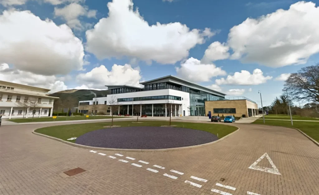 A modern veterinary school building, presenting a sleek combination of glass and concrete architecture. A rounded glass facade adjoins a more traditional block, reflecting a clear sky. The school's name is prominently displayed, asserting its institutional pride. Behind, lush greenery and rolling hills paint a serene backdrop, suggesting a harmonious blend of advanced education and natural beauty