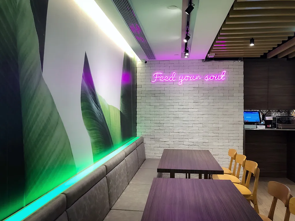 M Kitchen's vibrant dining atmosphere highlighted by neon 'Feed your soul' sign, large leaf murals, and sleek seating, an experience crafted by Rebel Arkitekt.