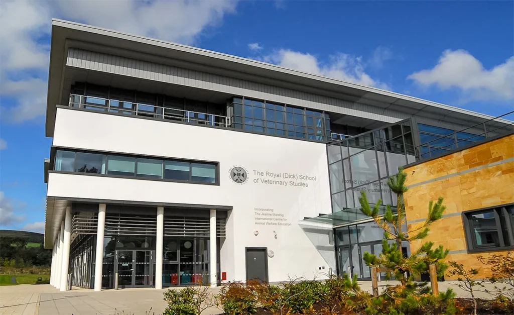 Front entrance of the Royal (Dick) School of Veterinary Studies showing a modern design with white and glass facades and stone cladding. Manicured lawn with young conifers in front and the school's name displayed above the entrance, with the Jeanne Marchig International Centre for Animal Welfare Education mentioned. Clear sky and lush greenery in the background.