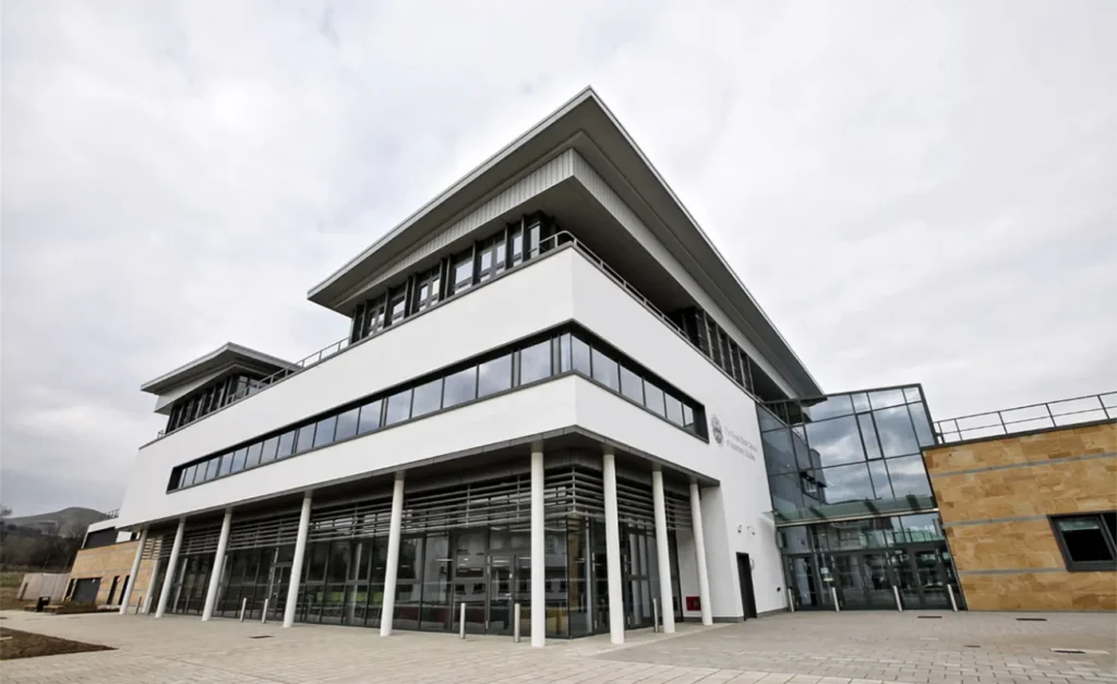 Angular view of a modern veterinary school building featuring a mix of glass, white panels, and stone cladding. A prominent glass atrium intersects the structure, and the foreground displays a paved approach leading to the main entrance, with a cloudy sky overhead.