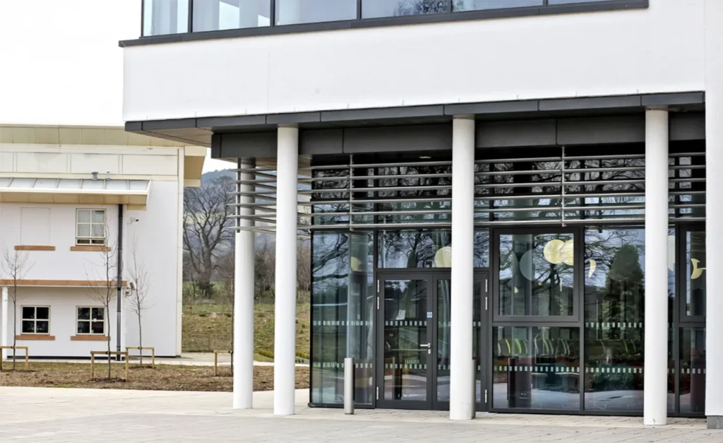 Frontal view of a contemporary veterinary school building, featuring a contrasting blend of white and sandy-hued stone facades. The entrance is framed by a row of slender white columns and expansive glass panels, allowing a glimpse of the interior.