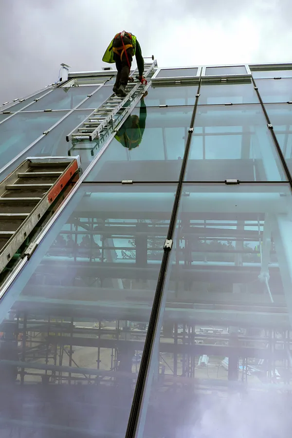 Worker on a ladder against a large angled glass structure during construction.