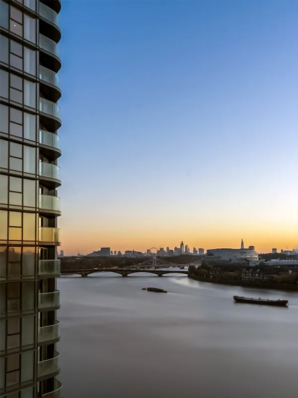 Dawn breaks over London, with the silhouette of a modern skyscraper on the left and the serene River Thames flowing towards a distant city skyline, featuring the silhouettes of iconic buildings under a clear gradient sky.