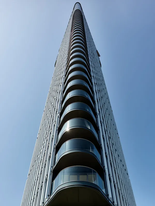 Looking up at a modern skyscraper with a pattern of rounded balconies, the facade is adorned with reflective glass that mirrors the clear blue sky, emphasizing the building's towering presence and architectural elegance.