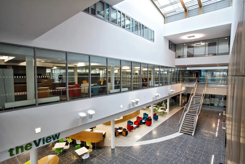 Spacious double-height atrium inside a veterinary school, featuring a library visible through large glass windows, modern seating areas, and a central staircase connecting the floors.