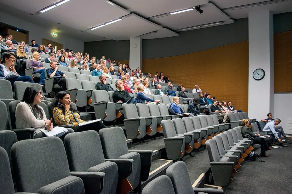 A lecture hall filled with attentive students seated in tiered, grey upholstered chairs, engaging with a presentation in a contemporary veterinary school setting, with warm lighting and a clock on the back wall indicating the time.