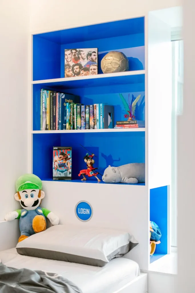 Cheerful children's bedroom shelving unit, brought to life with vibrant blue colors and a collection of books and toys, complemented by a plush character toy on the bed, designed by Rebel Arkitekt for a personalized touch.