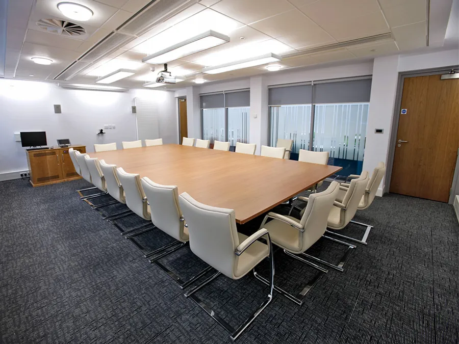 Spacious conference room setup with large wooden table, cream chairs, and modern audiovisual equipment in the background.