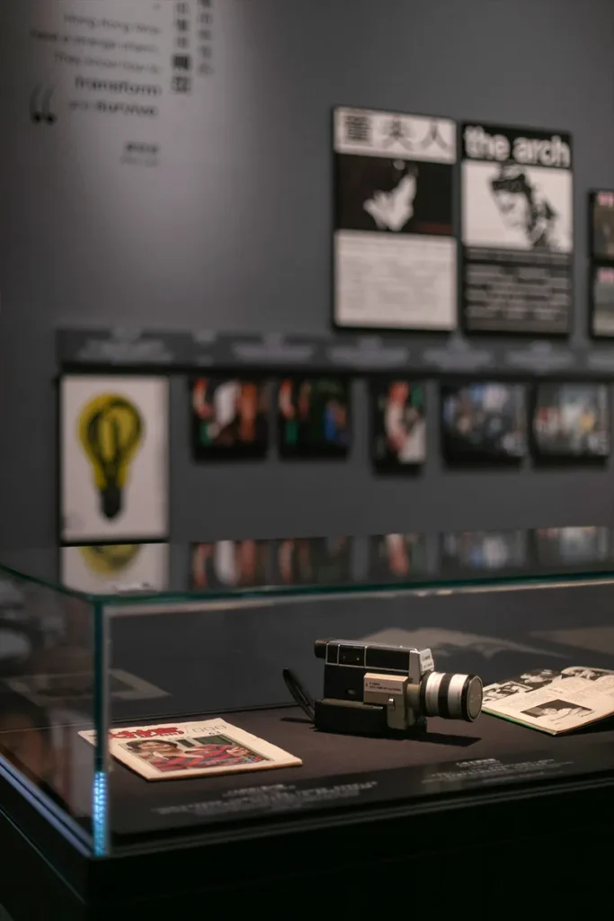 Vintage movie camera on display at a museum exhibition, with focus on the device placed in a glass case against a backdrop of iconic film quotes and monochrome promotional movie stills.