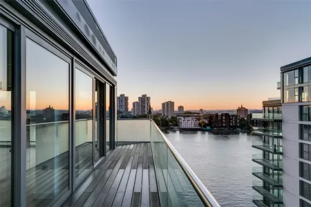 Sunset view from a waterfront apartment balcony, capturing the reflective glass railing and the tranquil waters of the Thames with the London cityscape unfolding in the distance, as the sky transitions to evening with gradients of orange and blue.