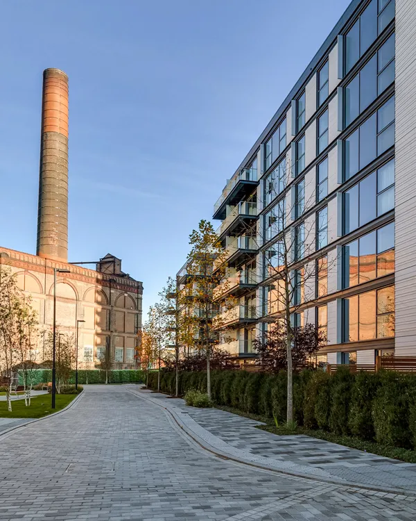 The late afternoon sun illuminates the back elevation of a waterfront building, highlighting its modern architecture and reflective glass windows, set against the backdrop of the historic Lots Road Power Station, with a neatly paved walkway leading through a lush, landscaped garden.