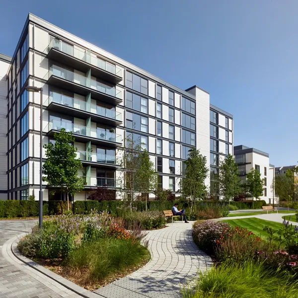 A person enjoys a quiet moment on a bench in a landscaped communal garden of a contemporary residential development, featuring a curving pathway, lush plantings, and the building's modern façade with expansive balconies and clean lines against a clear sky.