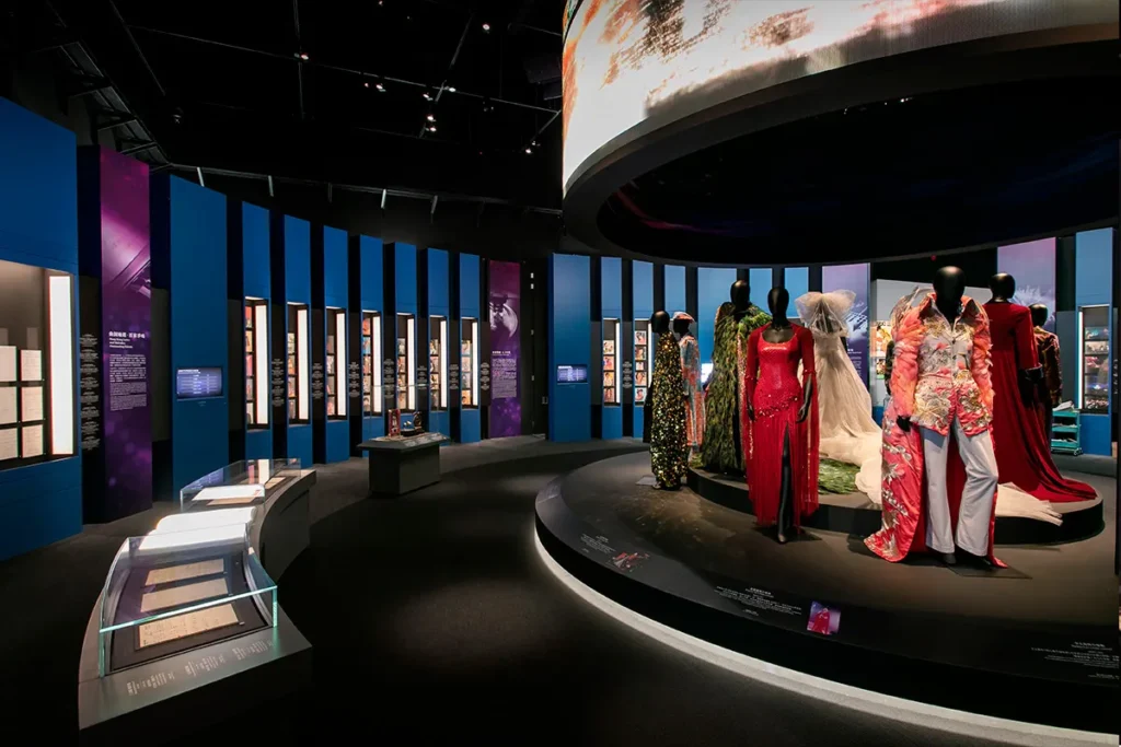 The exhibit features a central circular stage showcasing a collection of exquisite costumes, surrounded by blue vertical panels with informational content, highlighting Rebel Arkitekt's project management in bringing to life the theatrical heritage as part of Hong Kong's vibrant cultural scene.