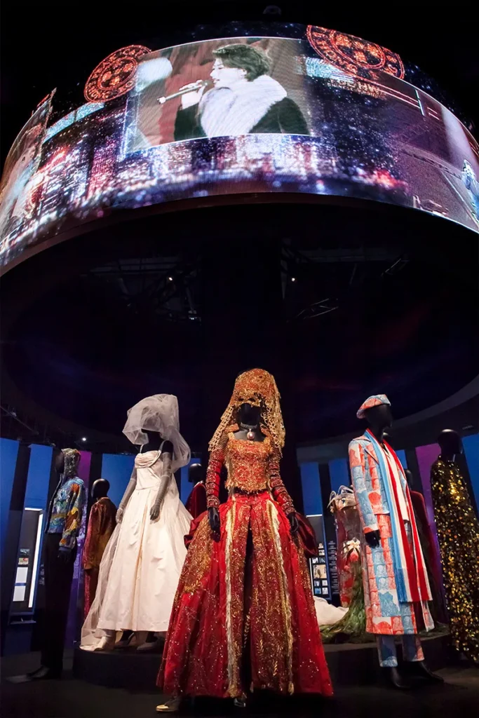 A circular stage surrounded by theatrical costumes ranging from traditional to extravagant; above, an LED screen projects vivid images, creating an immersive atmosphere of performance and design.