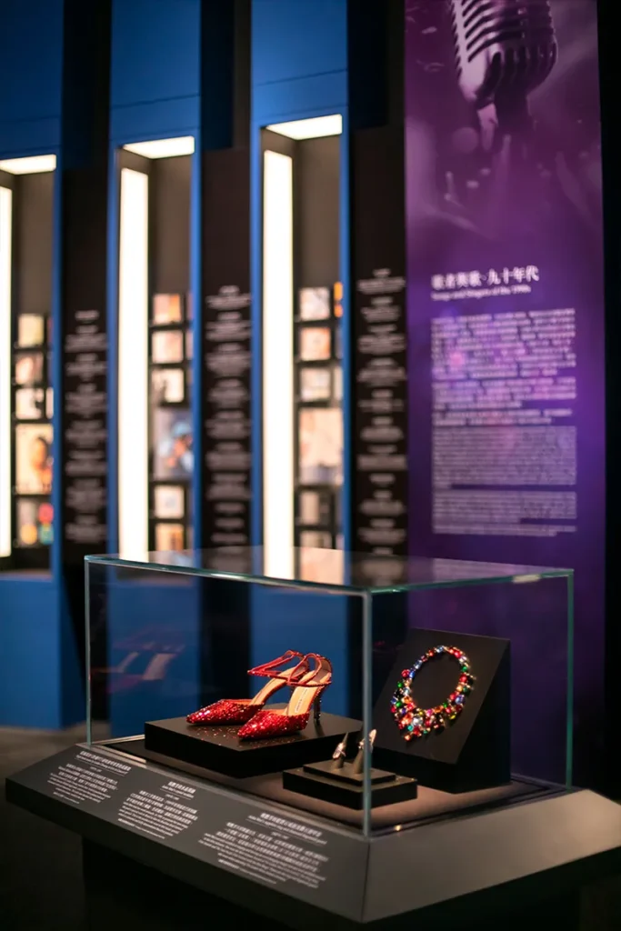 Elegant red high-heel shoes and a colorful necklace displayed in a glass case, with informative plaques, against a backdrop of vertical panels with mounted frames, and a large purple banner with text.