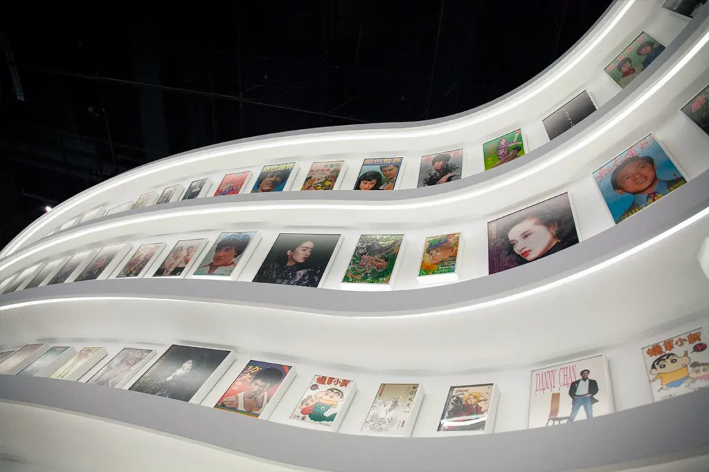 Curved, ascending white shelves showcase a variety of classic film and music album covers, tracing the evolution of Hong Kong's pop culture and entertainment