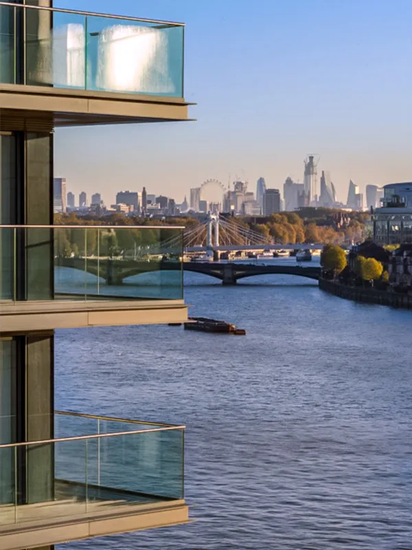 Partial view of apartment balconies overlooking the River Thames with London's skyline in the distance, including the London Eye and a barge floating by, all under a clear sky at dusk, highlighting the urban backdrop against the calmness of the river.