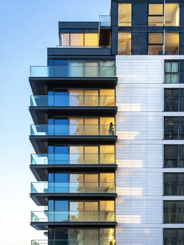 The evening sun reflects off the glass balconies of a modern apartment building, showcasing the clean lines and reflective surfaces of its architecture, with a clear blue sky in the background creating a contrast that highlights the building’s urban design.